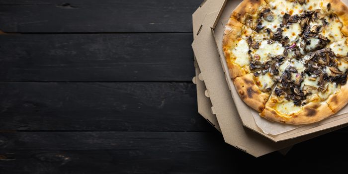 Pizza funghi in an open delivery box, dark backdrop with text space. Delivery foods, takeaway food concept: pile of mushroom pizzas in boxes on black wood background