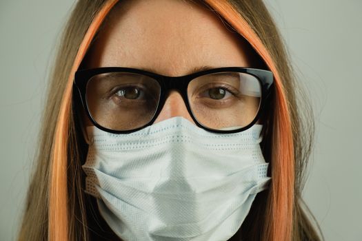 Intelligent woman in eyeglasses wearing medical mask, close-up portrait with focus on eyes. Young female in surgical mask in dark gray backdrop. Concept of personal hygiene, protection against virus and germs or professional gear