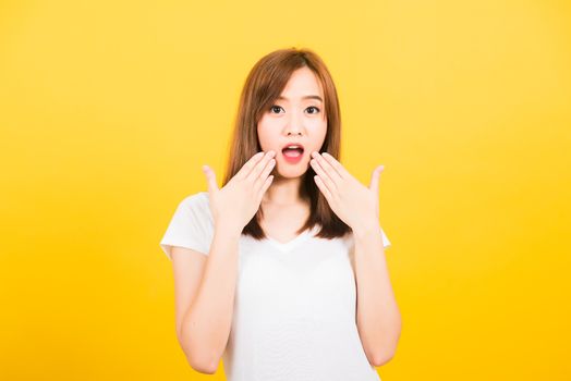 Asian happy portrait beautiful cute young woman teen standing amazed, shocked afraid wide open mouth eyes gesturing palms looking to camera isolated, studio shot on yellow background with copy space