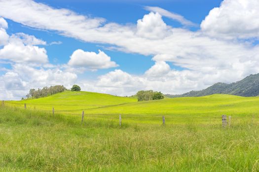 Australian country landscape scenic background showing a barbed wire fence on rural agricultural pasture with green grass and cloudy blue sky and sunlit hills