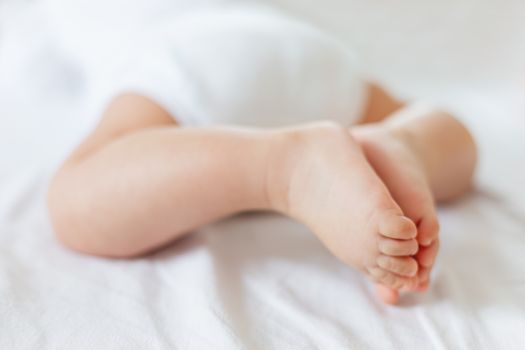 Baby's feet. Bare heels of little child wearing white bodysuit and diaper. Cozy morning bedtime at home.