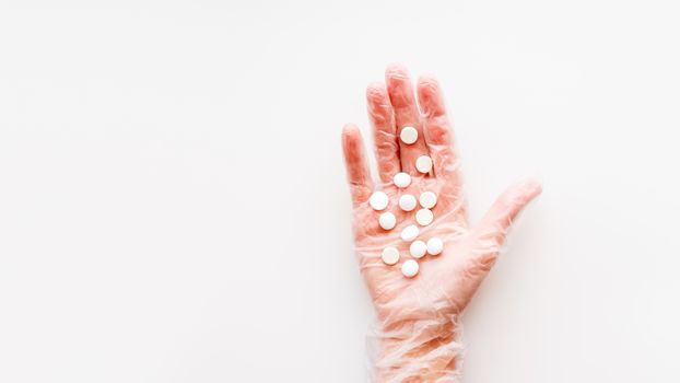 Doctor's palm hand in protective transparent glove full of white scattering pills. Capsules with medicines on white background with copy space. Flat lay, top view.