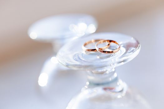 Top view on golden wedding rings lying on top of upside down staying wine glasses. Sun shines through transparent glass.