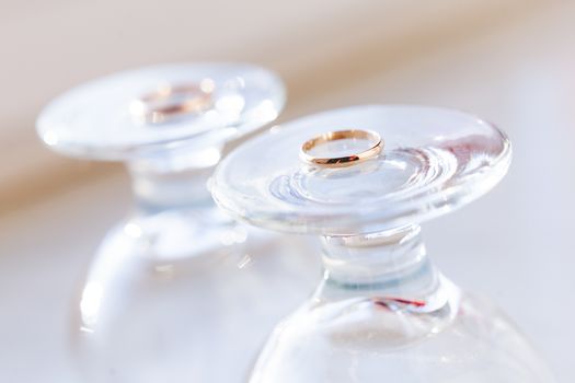 Top view on golden wedding rings lying on top of upside down staying wine glasses. Sun shines through transparent glass.