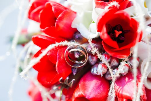 Golden wedding rings on bridal bouquet. Symbol of love and marriage on floral composition of bright red tulips.