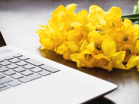 Bouquet of Narcissus or daffodils lying on silver metal laptop. Bright yellow flowers on portable device. Wooden background.