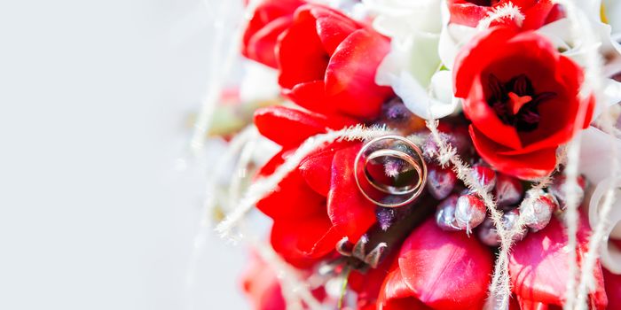 Golden wedding rings on bridal bouquet. Symbol of love and marriage on floral composition of bright red tulips. Background with copy space.