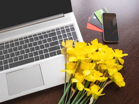 Bouquet of Narcissus or daffodils lying on silver metal laptop. Top view on bright yellow flowers on portable device and smartphone with credit cards on wooden table.