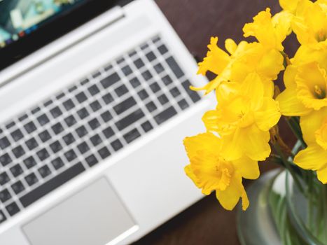 Bouquet of Narcissus or daffodils in glass vase over silver metal laptop. Bright yellow flowers with portable device. Wooden background.