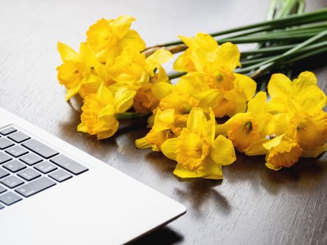 Bouquet of Narcissus or daffodils lying on silver metal laptop. Bright yellow flowers on portable device. Wooden background.