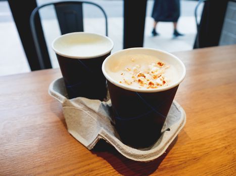 Two paper cups with tasty coffee on wooden table in cafe.. Hot beverage with popcorn on frothy milk. Holder made from recycled cardboard for both cups.