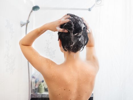 Naked woman with short hair takes a shower. Woman washes her hair with shampoo. White bathroom.