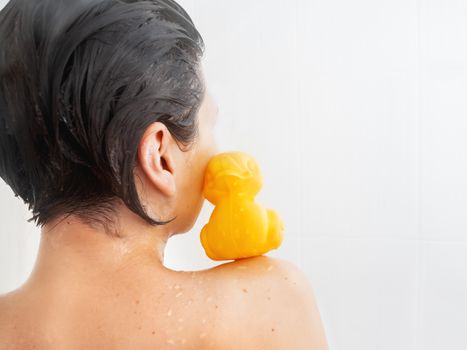 Naked woman with short hair takes a shower. Yellow rubber duck is sitting on her shoulder. Girl with funny toy in white bathroom.
