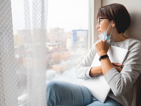 Woman in medical mask had lost her job. She sits on window sill with closed laptop. Lockdown quarantine because of coronavirus COVID19. Self isolation at home.