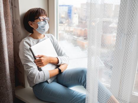 Woman in medical mask had lost her job. She sits on window sill with closed laptop. Lockdown quarantine because of coronavirus COVID19. Self isolation at home.