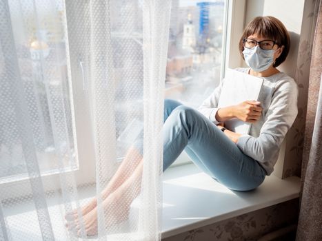 Woman in medical mask had lost her job. She sits on window sill with closed laptop. Lockdown quarantine because of coronavirus COVID19. Self isolation at home.