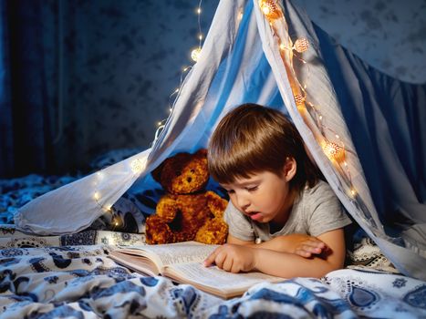 Little boy reads book. Toddler plays in tent made of linen sheet on bed. Cozy evening with favorite book.