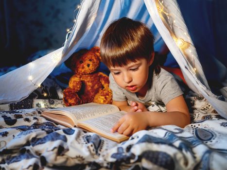 Little boy reads book with his teddy bear. Toddler plays in tent made of linen sheet on bed. Cozy evening with favorite book.