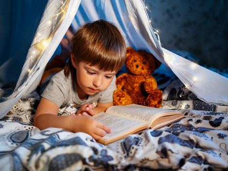 Little boy reads book with his teddy bear. Toddler plays in tent made of linen sheet on bed. Cozy evening with favorite book.