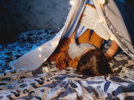 Little boy reads book. Toddler plays in tent made of linen sheet on bed. Cozy evening with favorite book.