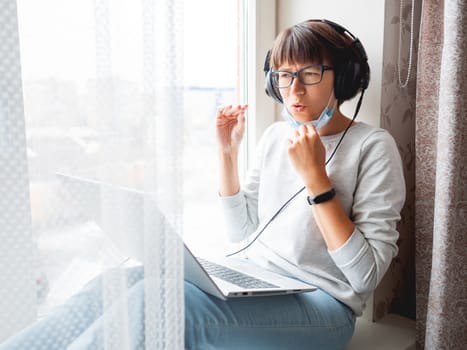 Woman in medical mask remote works from home. She speaks online with somebody on window sill with laptop on knees. Lockdown quarantine because of coronavirus COVID19. Self isolation at home.