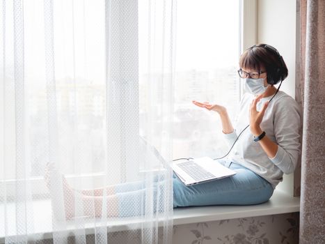 Woman in medical mask remote works from home. She speaks online with somebody on window sill with laptop on knees. Lockdown quarantine because of coronavirus COVID19. Self isolation at home.