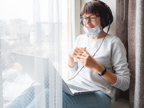 Woman in medical mask remote works from home. She speaks online with somebody on window sill with laptop on knees. Lockdown quarantine because of coronavirus COVID19. Self isolation at home.