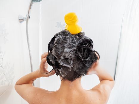 Naked woman with short hair takes a shower. Woman washes her hair with shampoo. Yellow rubber duck on girl's head in white bathroom.