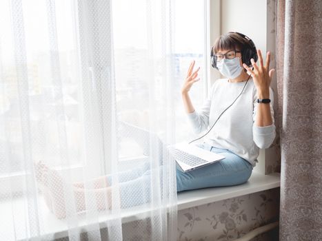 Woman in medical mask remote works from home. She speaks online with somebody on window sill with laptop on knees. Lockdown quarantine because of coronavirus COVID19. Self isolation at home.