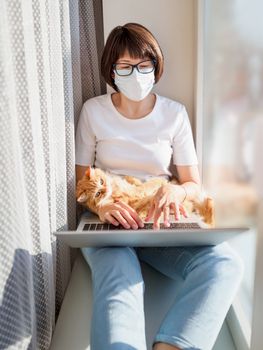 Woman in medical mask remote works from home. She sits on window sill with laptop and cute ginger cat on her knees. Lockdown quarantine because of coronavirus COVID19. Self isolation at home.