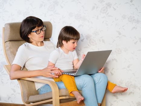 Mother and her toddler boy sit together on chair with laptop. Woman tries to remote work, but kid is asking for game or cartoons. quarantine lockdown because of coronavirus COVID-19.