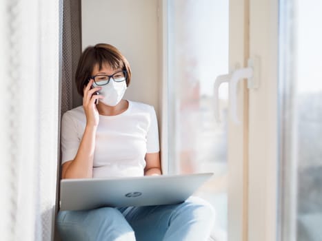 Woman in medical mask remote works from home. She sits on window sill with smartphone and laptop on knees. Lockdown quarantine because of coronavirus COVID19. Self isolation at home.