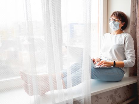 Woman in medical mask remote works from home. She sits on window sill with laptop on knees. Lockdown quarantine because of coronavirus COVID19. Self isolation at home.