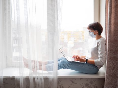 Woman in medical mask remote works from home. She sits on window sill with laptop on knees. Lockdown quarantine because of coronavirus COVID19. Self isolation at home.