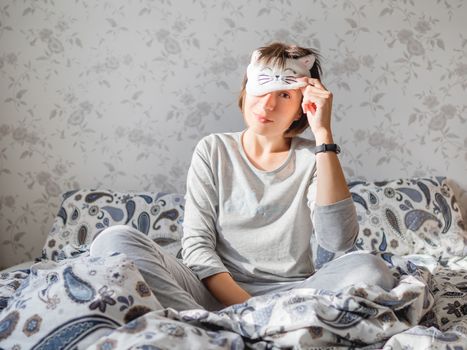 Young woman in grey pajama and sleeping mask in shape of cute sleeping cat face. She is just woke up and sit in bed. Early morning in cozy home.