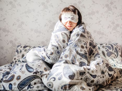 Young woman in grey pajama and sleeping mask in shape of cute sleeping cat face. She is just woke up and sit in bed. Early morning in cozy home.