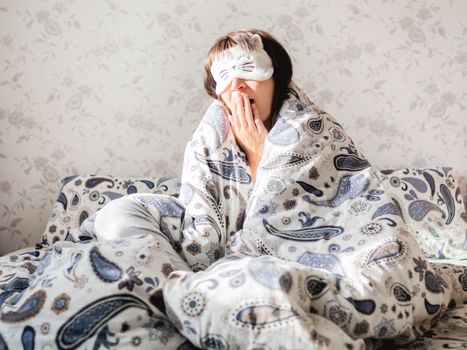 Young woman in grey pajama and sleeping mask in shape of cute sleeping cat face. She is just woke up and sit in bed. Early morning in cozy home.