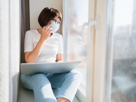 Woman in medical mask remote works from home. She sits on window sill with smartphone and laptop on knees. Lockdown quarantine because of coronavirus COVID19. Self isolation at home.