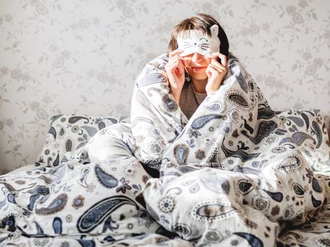 Young woman in grey pajama and sleeping mask in shape of cute sleeping cat face. She is just woke up and sit in bed. Early morning in cozy home.