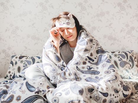 Young woman in grey pajama and sleeping mask in shape of cute sleeping cat face. She is just woke up and sit in bed. Early morning in cozy home.