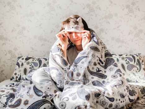 Young woman in grey pajama and sleeping mask in shape of cute sleeping cat face. She is just woke up and sit in bed. Early morning in cozy home.