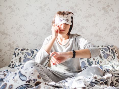 Young woman in grey pajama and sleeping mask in shape of cute sleeping cat face. She is just woke up and sit in bed. Early morning in cozy home.