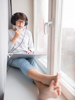 Woman in medical mask remote works from home. She sits on window sill with laptop on knees and headphones. Lockdown quarantine because of coronavirus COVID19. Self isolation at home.