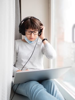 Woman in medical mask remote works from home. She sits on window sill with laptop on knees and headphones. Lockdown quarantine because of coronavirus COVID19. Self isolation at home.