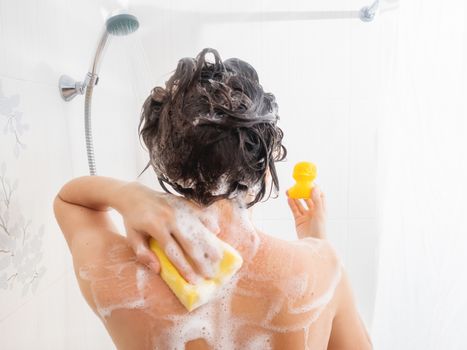 Naked woman with short hair takes a shower. Woman washes her hair with shampoo. Yellow rubber duck on girl's palm in white bathroom.