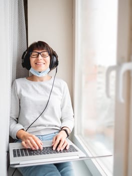 Woman in medical mask remote works from home. She sits on window sill with laptop on knees and headphones. Lockdown quarantine because of coronavirus COVID19. Self isolation at home.