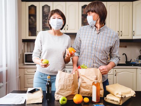 Young couple in protective masks is sorting out purchases in the kitchen. Products in bags made of craft paper. Food delivery in conditions of quarantine because of coronavirus COVID19.