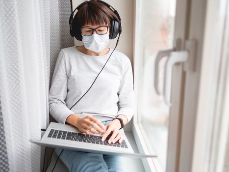 Woman in medical mask remote works from home. She sits on window sill with laptop on knees and headphones. Lockdown quarantine because of coronavirus COVID19. Self isolation at home.