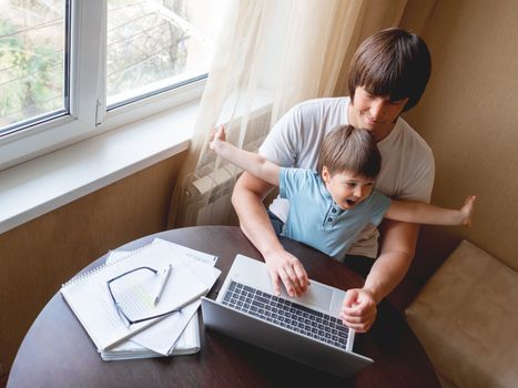 Self-isolation because of coronavirus outbreak. Man is remote working with his little son. Laptop on table.