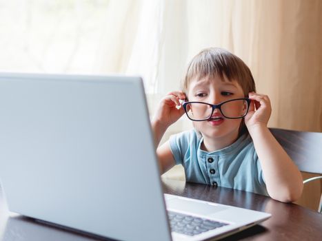 Curious toddler boy explores the laptop and staring on screen through big glasses. Symbol of online education for toddlers or impaired vision from prolonged exposure to screens of electronic devices.